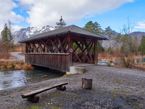 &Uuml;berdachte Holzbr&uuml;cke &uuml;ber einen Flussarm vor schneebedeckten Alpengipfeln