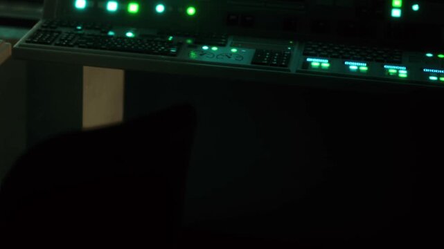 A control room shows a desk with several keyboards and buttons lit up in green. The scene takes place at night in a dark environment. The lights create a high tech feel.