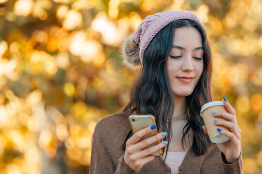 Young woman in a pink beanie enjoying autumn, smiling while holding a coffee cup and using a smartphone outdoors with golden fall foliage in the background