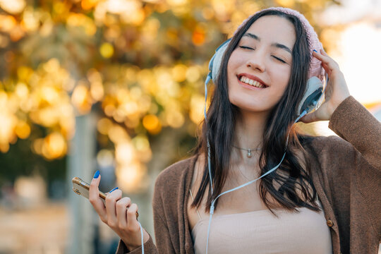 young woman smiling and listening to music with headphones, holding a smartphone, autumn park background with golden bokeh leaves