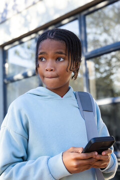African male teen standing before glass wearing light blue hoodie strap holding phone looking left