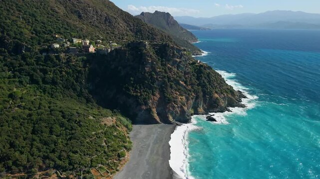 Nonza Cap Corse Corsica 4K drone video. Backward fly out reveal of vertical cliff village with long empty black sand beach and foaming turquoise sea waves. Scenic  travel b-roll cinematic footage