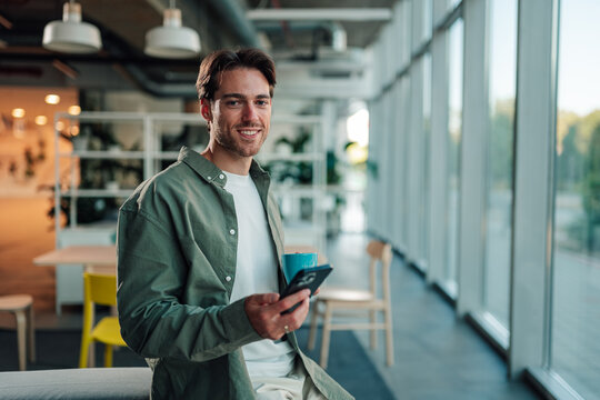 Young man smiling, holding smartphone and coffee mug, standing in a modern office lounge area