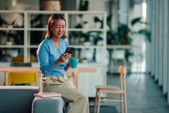 Young asian woman holding mobile phone and coffee cup, sitting on office couch, engaging with social media or work