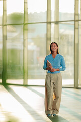 Fototapeta premium Asian woman standing and smiling, holding a notebook in a modern office with glass walls and natural light, depicting business success