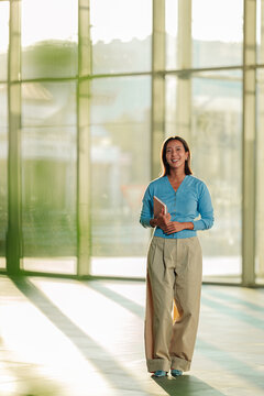 Asian woman standing and smiling, holding a notebook in a modern office with glass walls and natural light, depicting business success