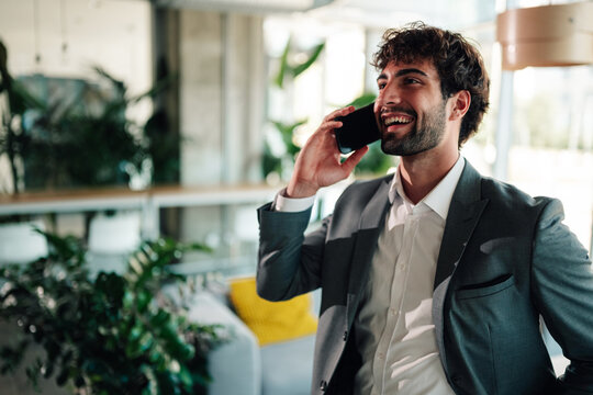 Young businessman talking on phone, smiling broadly. Professional man communicating in a bright office environment