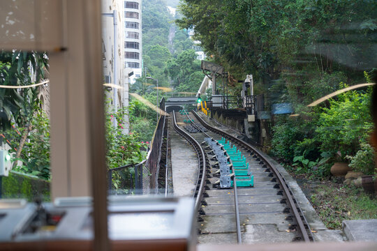 View from inside a funicular cabin traveling up a steep incline, passing a concrete station platform and tropical vegetation under soft daylight.