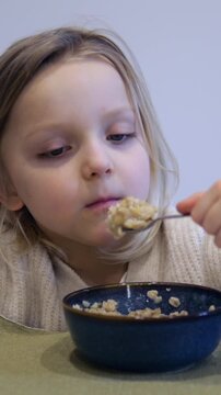 Adorable little blonde girl eating a bowl of oatmeal for breakfast, making a funny disgusted face because she dislikes the taste of the healthy food, showcasing a picky eater's reaction