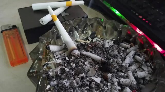Burning Cigarette in Full Glass Ashtray with Ashes Smoke Lighter and Keyboard Background