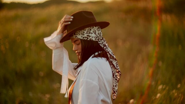 Young woman with patterned headscarf standing in a quiet grassy meadow during golden hour sunset