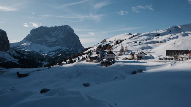 Snow blankets mountain peaks under bright blue sky. Sunlight glints on pristine white slopes. Village buildings dot the snowy valley floor. Mountain range forms a majestic backdrop