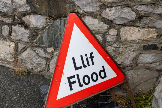 Weathered bilingual Lif Flood warning sign at Penrhyn Bay Wales displayed beside a rough stone wall with bold red and white triangular design and clear Welsh and English wording