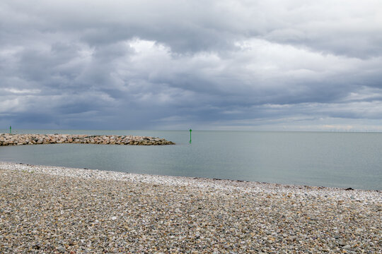 Pebble beach and calm sea at Penrhyn Bay Wales with rocky breakwater green navigation markers distant offshore wind turbines and a broad dramatic cloudy sky stretching over the coastline