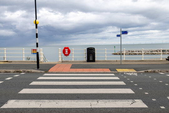 Pedestrian zebra crossing beside the seafront at Penrhyn Bay Wales with Belisha beacon cycle lane railings lifebuoy litter bin rocky breakwater and calm sea beneath a dramatic cloudy sky