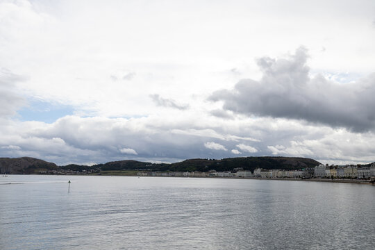 Wide view across Llandudno Bay in North Wales showing the curved Victorian seafront beneath dramatic clouds with surrounding hills and calm coastal water stretching across the foreground