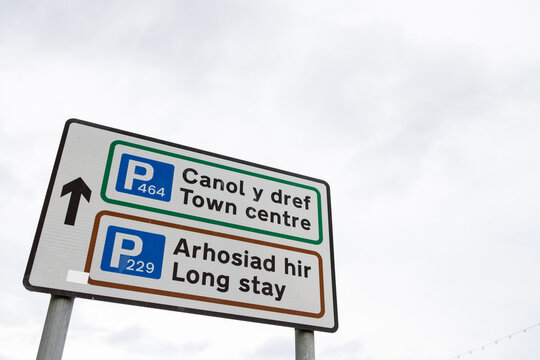 Bilingual parking direction sign in Llandudno North Wales showing town centre and long stay parking information in Welsh and English with route numbers and an upward arrow beneath cloudy sky