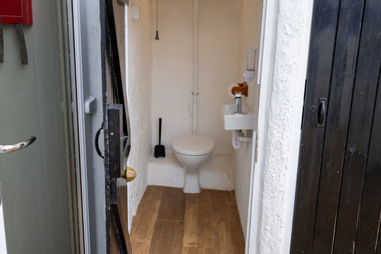 Outside toilet at Penrhyn Bay North Wales seen from the entrance with open dark door white WC compact hand basin wooden floor exposed pipes toilet brush and wall dispensers