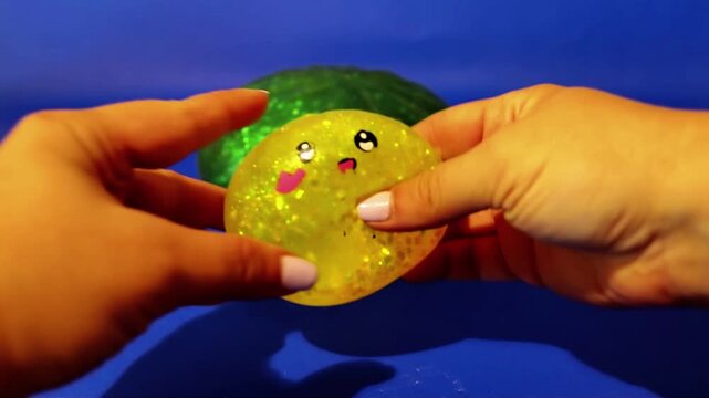 A hand squeezes a bright neon yellow glittery dumpling toy against a vibrant blue background, with colorful squishy toys nearby
