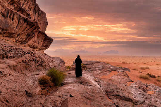 Rear view silhouette of a person standing on rocky outcrop watching a dramatic fiery golden sunset over the desert plain in Tabuk