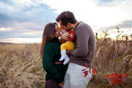 Young parents hugging infant baby girl walking outdoors in fall meadow. Happy loving family kissing daughter enjoying autumn view