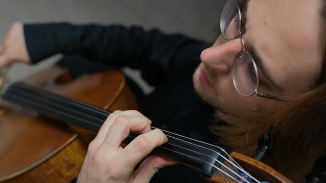 Caucasian young adult man with glasses playing cello, pressing fingers on strings in close up. Video is ideal for music school commercials, online learning platforms, or classical concert promos