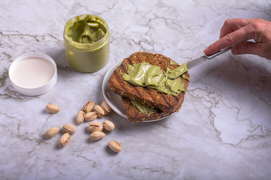 Two slices of toasted artisan bread spread with smooth pistachio cream and sprinkled with chopped nuts next to a glass jar and pestle