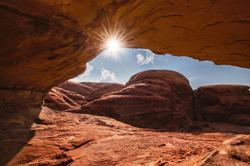 Wide shot of radiant sunburst shining through a natural sandstone cave arch over rugged red desert mountains in Tabuk © almozinisaleh