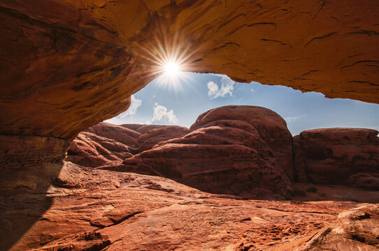 Wide shot of radiant sunburst shining through a natural sandstone cave arch over rugged red desert mountains in Tabuk