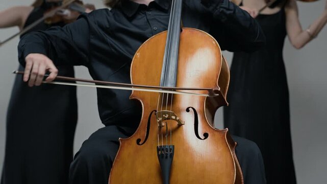 Young adult Caucasian male cellist playing instrument with bow, accompanied by female violinists in background. Clip is ideal for promoting classical music concerts or arts academies