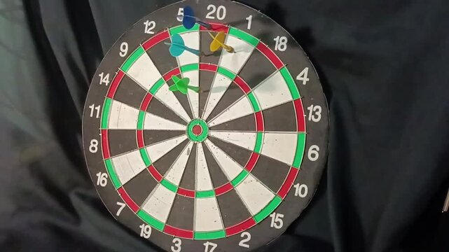 A close-up of a dartboard with several colorful darts stuck in the scoring sections, showing a recreational darts game setup 