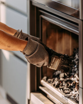 Indoor fireplace cleaning close up shows gloved hands using brush to remove soot from modern hearth, with warm light and uncluttered wall creating practical home maintenance scene