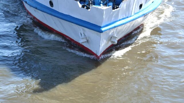 Bow of white and blue ship navigating river water, creating wake and shadows
