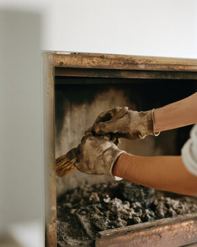 Indoor fireplace soot cleaning with gloved hands using handheld brush, showing careful maintenance work, dark ash residue, worn metal, warm room light, and clean wall