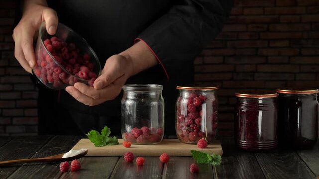 Chef in black uniform prepares fresh raspberries for canning, transferring them from a bowl into glass jars on a wooden board, with jars of jam and a rustic brick wall in the background