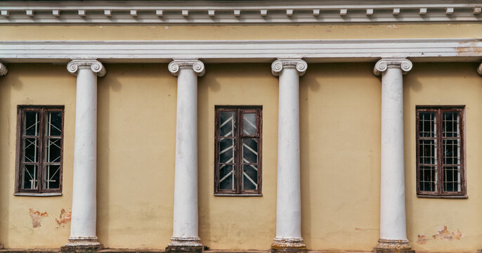 White ionic columns stand in a row along the yellow stucco facade of a historic building between old wooden framed windows.