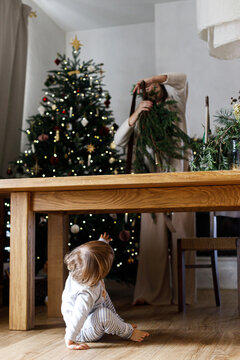 Toddler sitting under table while mother crafts evergreen garland near Christmas tree. Concept of cozy family holiday moments, domestic winter warmth, festive childhood curiosity.