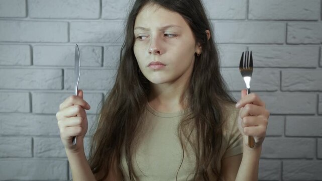 Sad hungry girl waiting for food holding cutlery. Unhappy young girl with dark circles under her eyes holding a fork and knife, patiently waiting for her meal, embodying the concept of hunger