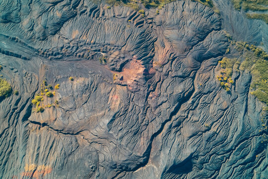 Eroded slag heap surface with deep ravines orange mineral deposits and patches of green, drone shot top view
