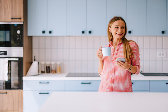 Smiling Woman in Modern Kitchen Holding Coffee and Smartphone, Casual Morning at Home