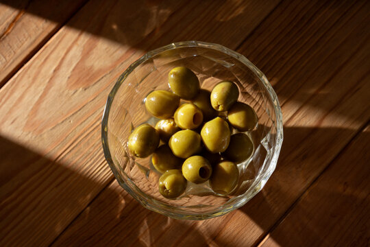 Green olives on a glass bowl lying on a wooden table with dramatic lateral harsh light, horizontal image