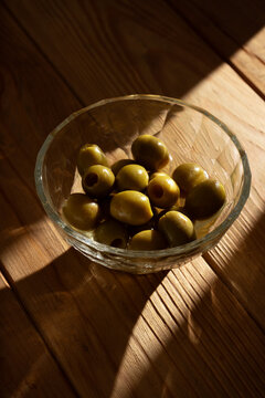 Green olives on a glass bowl lying on a wooden table with dramatic lateral harsh light, vertical image