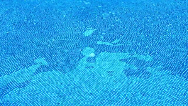 A swimming pool filled with clear blue water shows the reflection of sunlight on its surface. People enjoy swimming and playing in the water during the warm midday hours.
