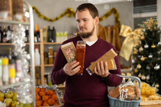 In festive little store decorated with Christmas tree and shiny tinsel, pensive young man with beard browsing shelves, picking out rich nutty turron and imagining joy of indulging in sweet delight