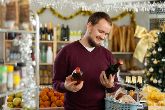 Man in a sweater buys whiskey at a Christmas supermarket. Customer chooses whiskey against the background of shelves with food decorated for the New Year