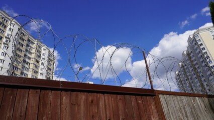 Wooden fence with barbed wire and apartments. A brown wooden fence topped with razor wire stands against blue sky. Tall apartment buildings rise in the background creating urban contrast. © nina blez