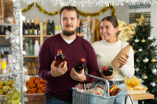 Couple with a basket of food choose alcohol for the New Year. Man and woman holding bottles of champagne and whiskey on the background of a Christmas tree