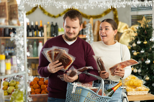 Married couple with a basket of groceries buys dried sausage and ham in supermarket - husband and wife are preparing to celebrate Christmas