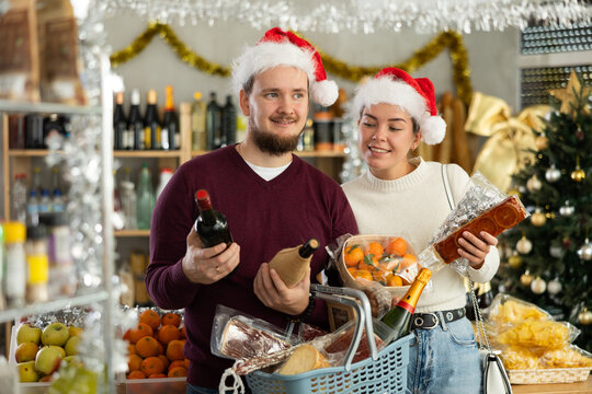 Couple in Santa hats holding tangerines, wine, sausage and basket of food in a Christmas supermarket. Man and woman shopping at a grocery store for the New Year
