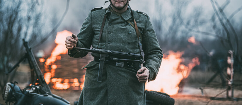 German Wehrmacht Infantry Soldier In World War Ii Standing Against The Background Of A Burning House. German WWII Soldier